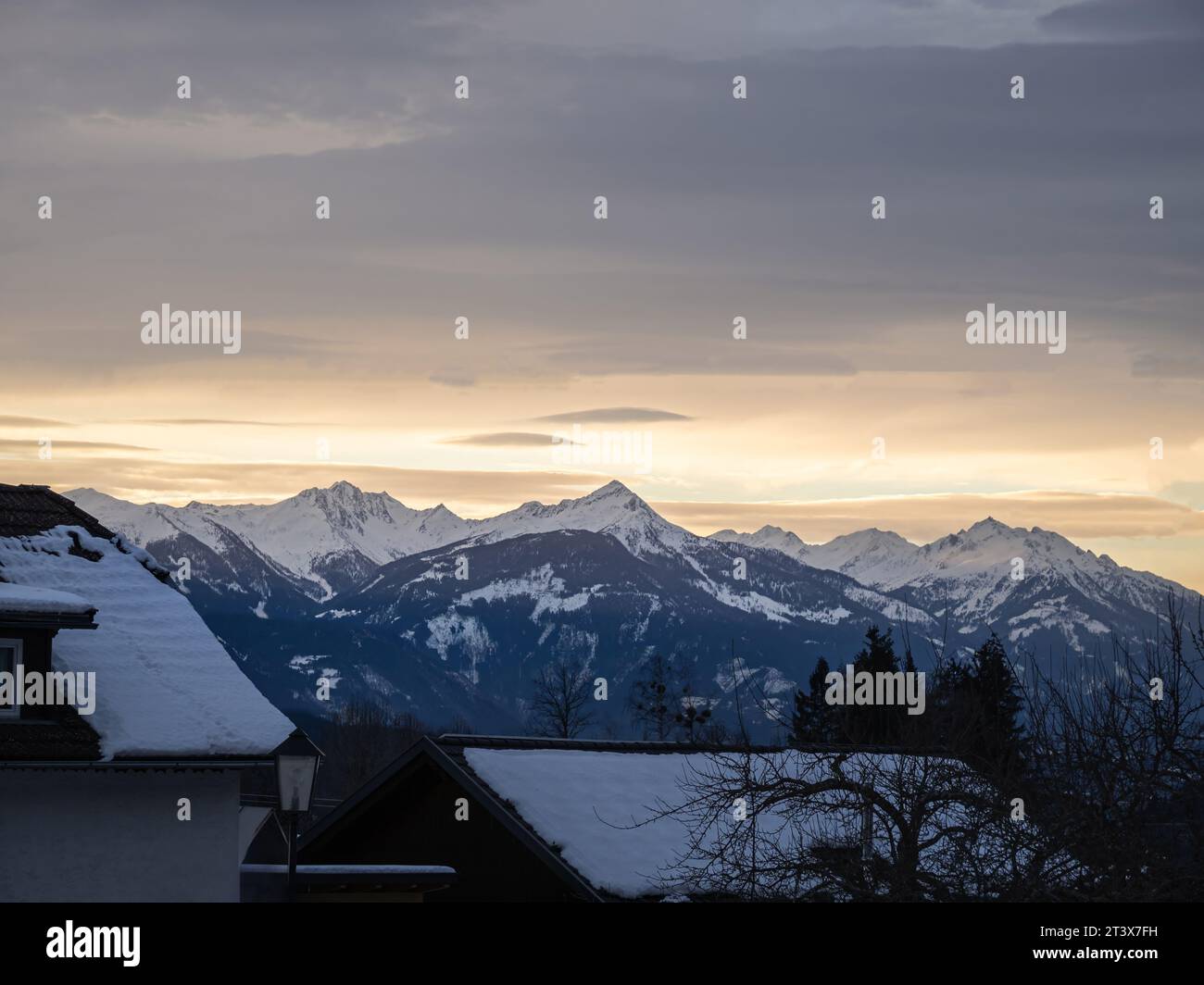 Puesta de sol colorida de la nieve del invierno sobre un pueblo de los alpes austriacos con las montañas en el fondo, tejados cubiertos de nieve, Carintia, Austria Foto de stock