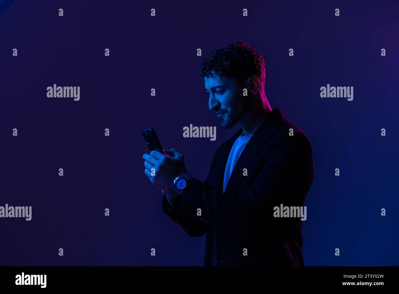 Hombre con el teléfono en la mano mirando la pantalla del smartphone, fondo azul oscuro del retrato, luz de neón, estilo y tendencias, luz mezclada, moda de los hombres, copiar pla Foto de stock