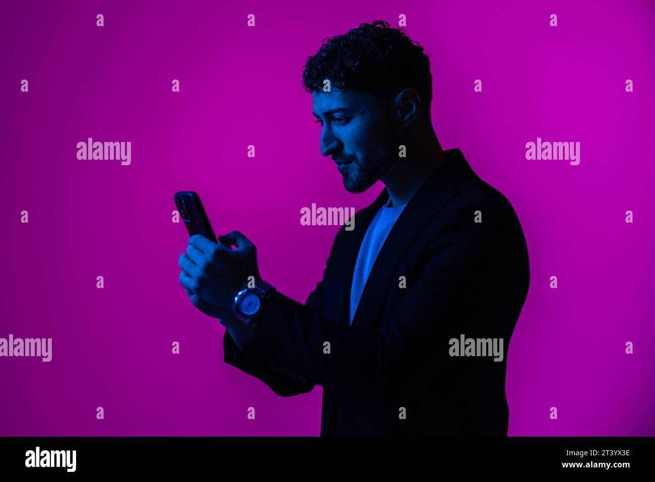 Hombre con el teléfono en la mano mirando la pantalla del smartphone, fondo azul oscuro del retrato, luz de neón, estilo y tendencias, luz mezclada, moda de los hombres, copiar pla Foto de stock