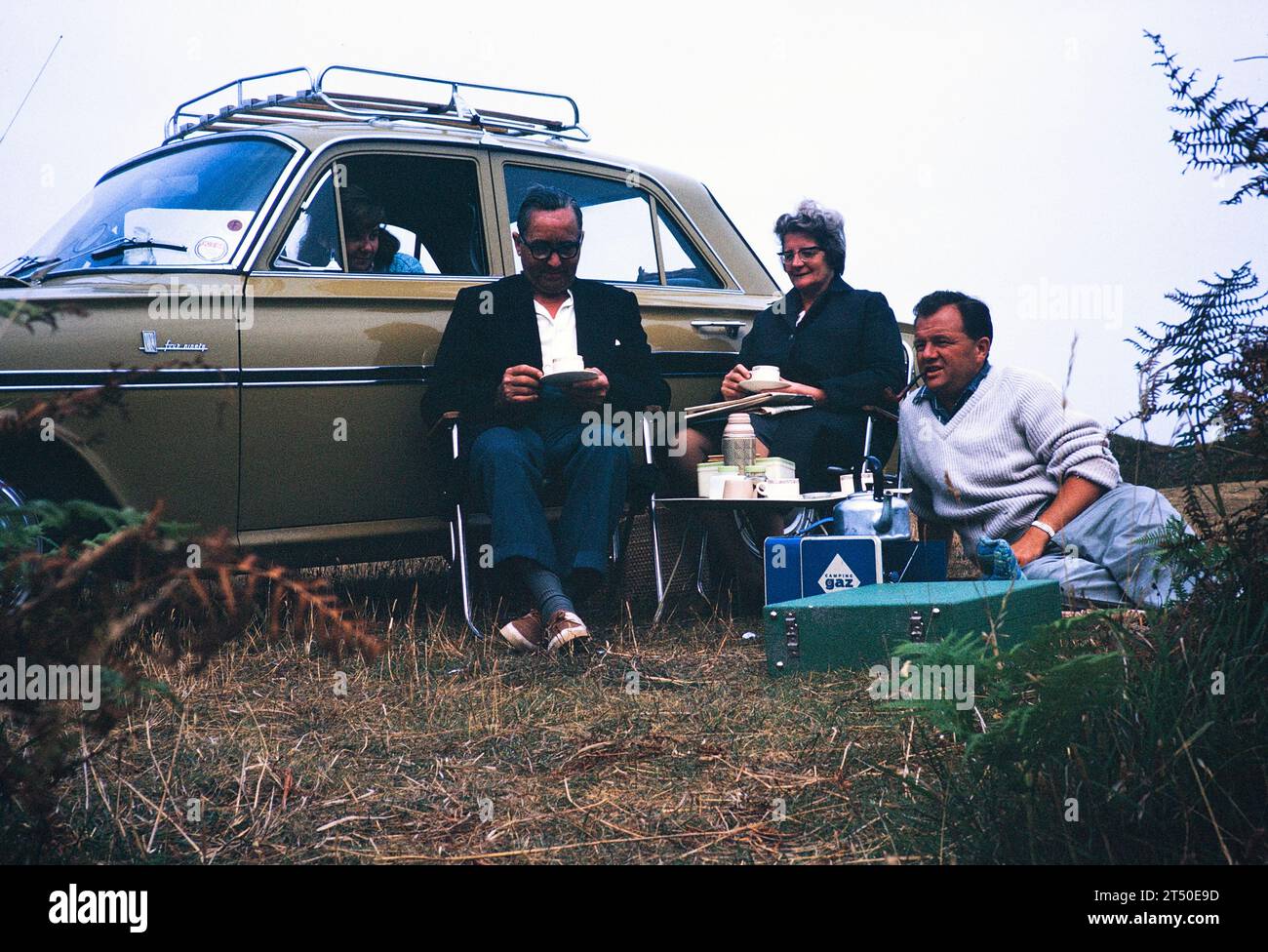 Picnic de la familia sentado en la estufa de gas de camping del coche, pipa de fumar del hombre, Reino Unido 1964 Foto de stock