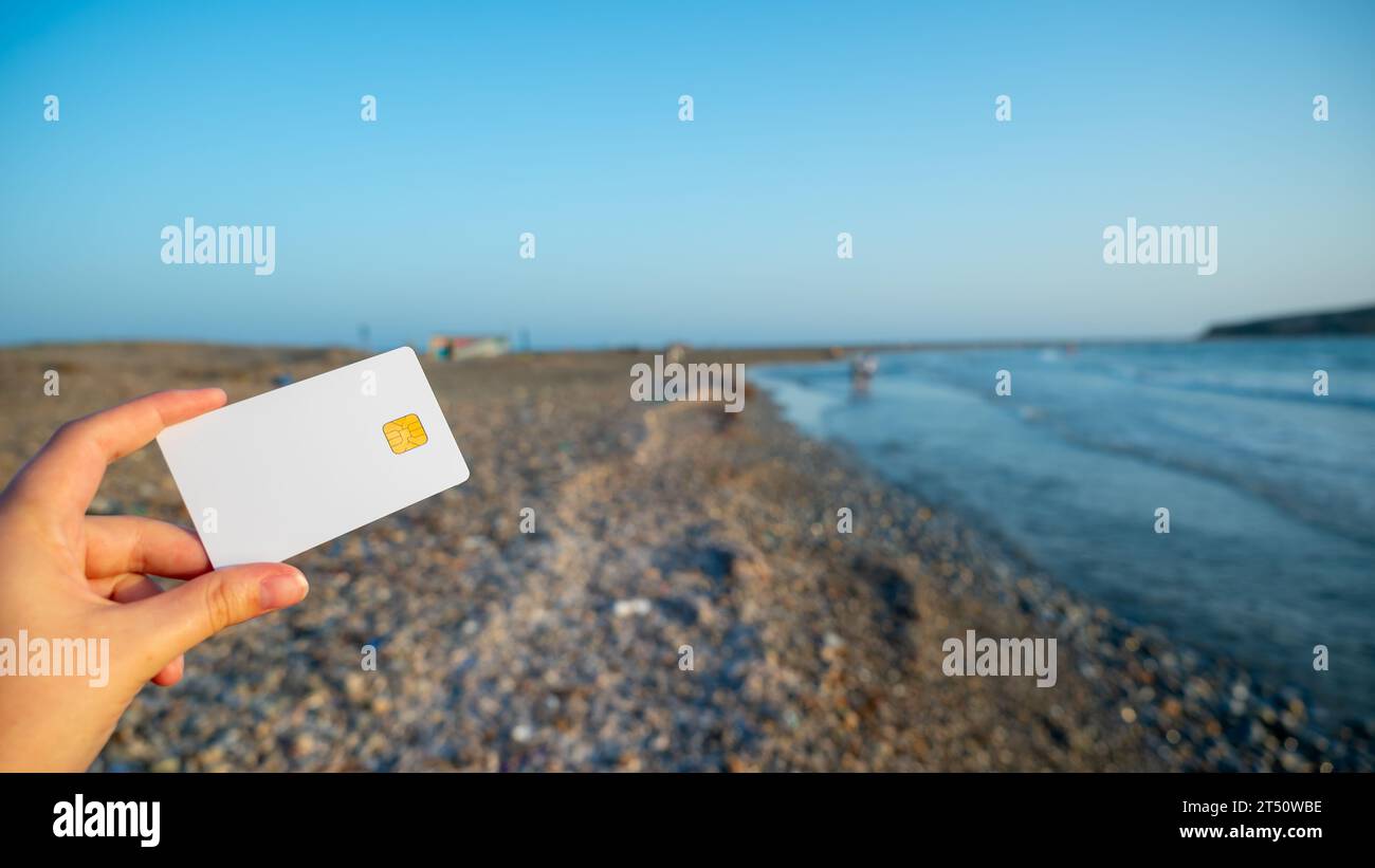 La mujer sostiene una tarjeta bancaria blanca en la mano contra el telón de fondo de una playa interminable como símbolo de posibilidades ilimitadas y viajes. copiar espacio Foto de stock