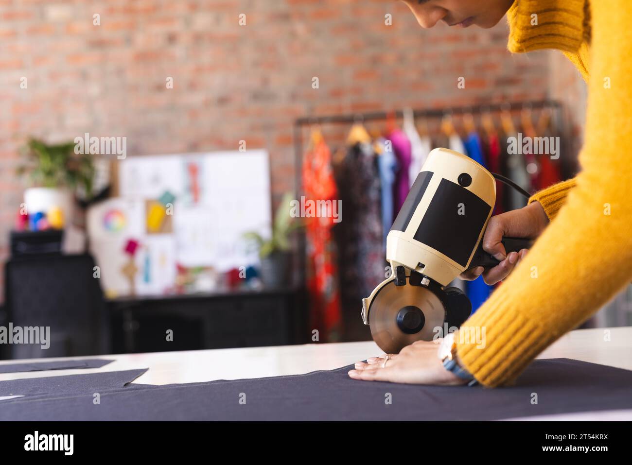 Diseñador de moda femenino biracial que usa un cortador de tela rotativo en un estudio soleado Foto de stock