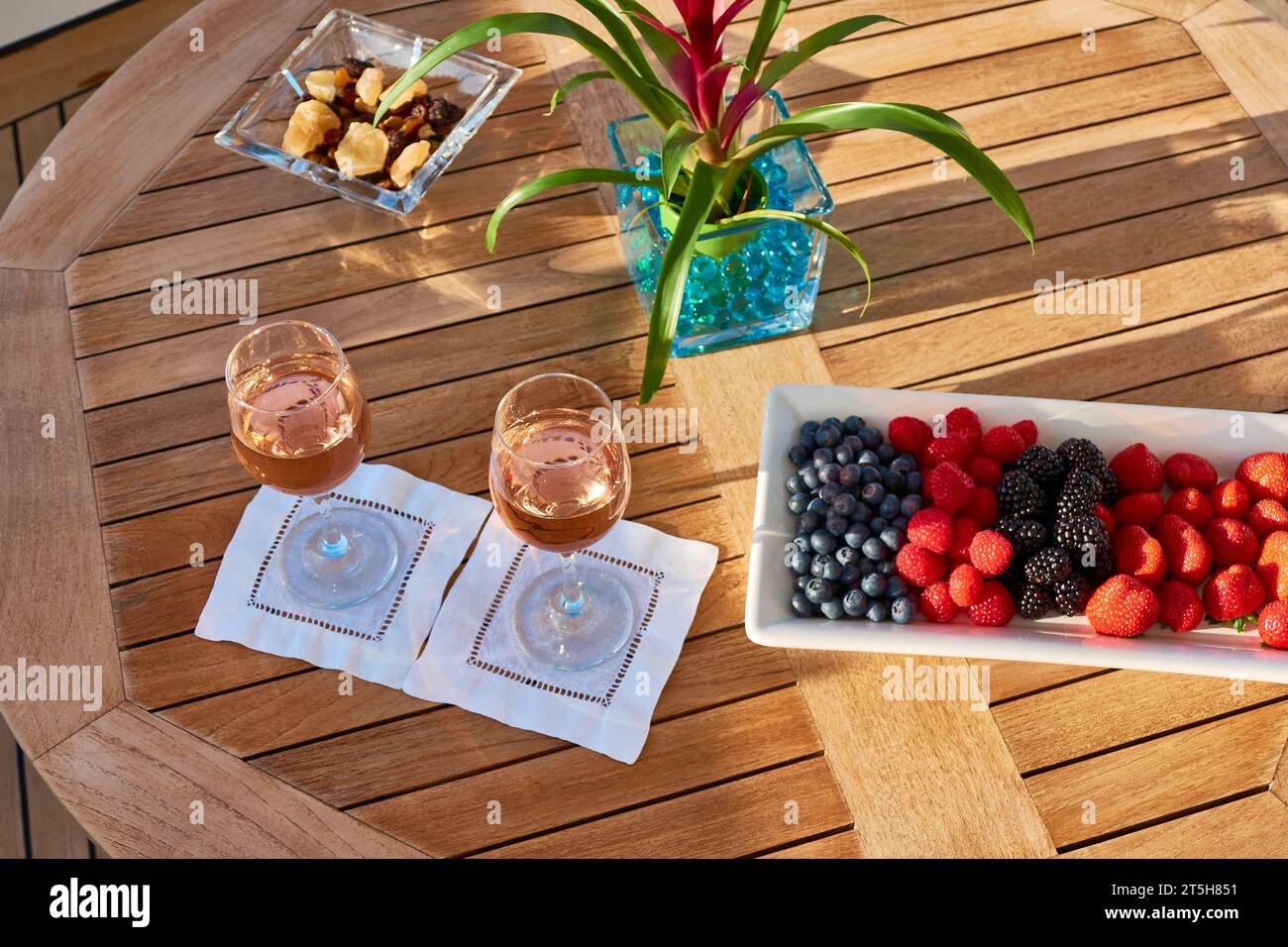 Dos copas de vino rosa en una mesa redonda de teca con un cactus floreciente y un tazón de arándanos, frambuesas y fresas. Foto de stock