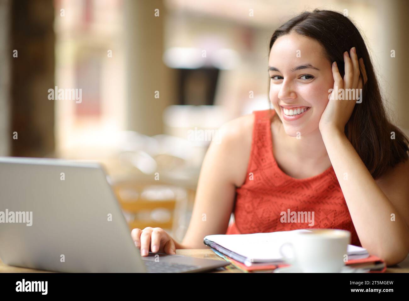 Estudiante feliz con el cuaderno y el ordenador portátil te mira en una terraza de la cafetería Foto de stock