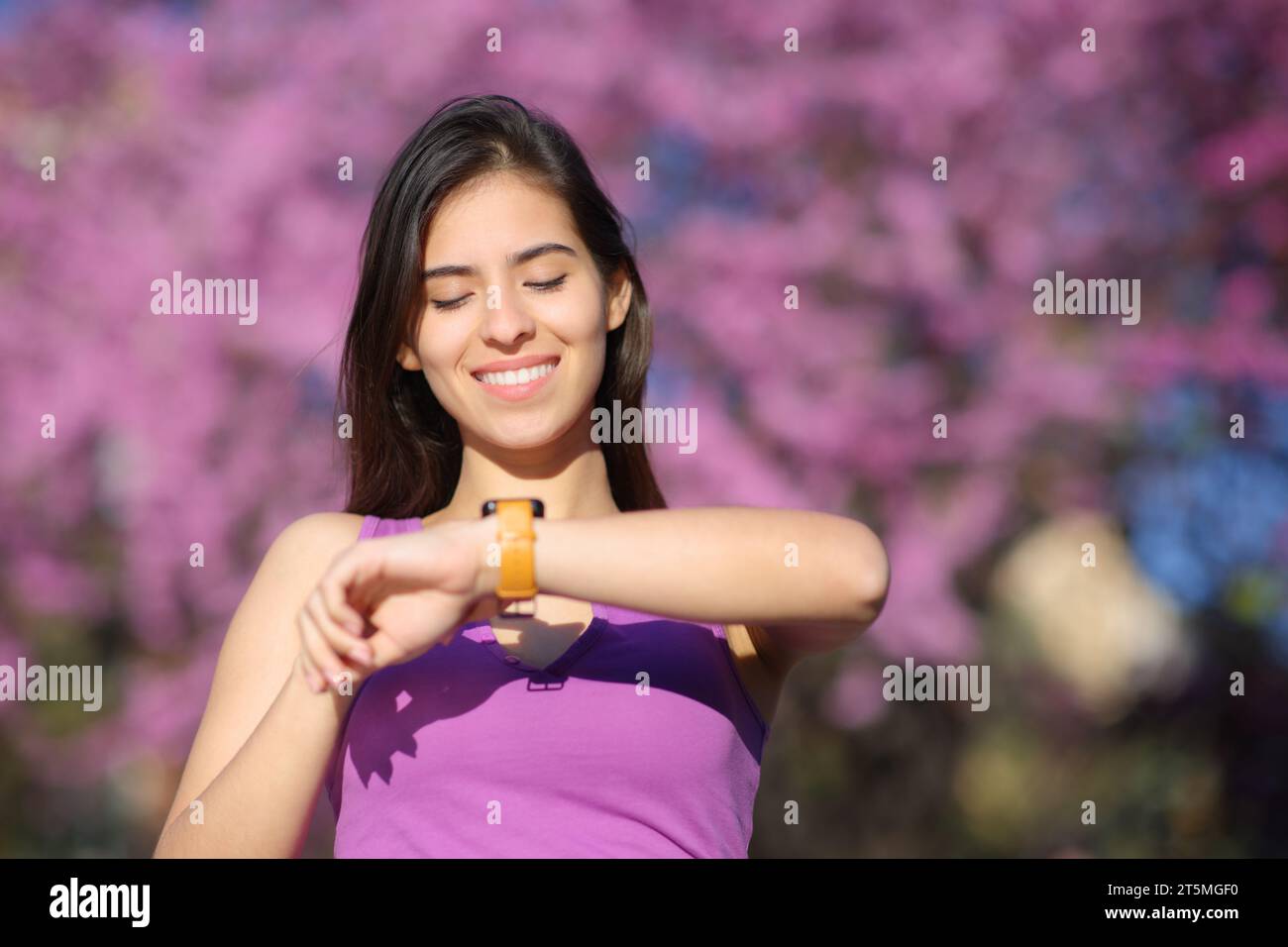 Retrato de la vista frontal de una mujer feliz que comprueba el smartwatch en un parque violeta Foto de stock