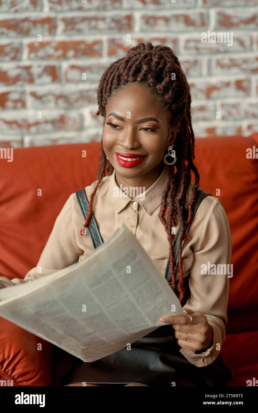 Bonita mujer africana leyendo el periódico y sonriendo. Señorita disfrutando de su tiempo de la mañana en casa, café u oficina. Artículo de historia o periódico de la mañana Foto de stock