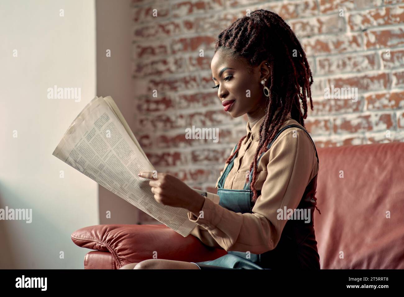 Atractiva mujer afroamericana con pavor peinado leyendo el periódico con interés mientras se sienta en un sofá de cuero rojo. La maldad de la gente de negocios Foto de stock