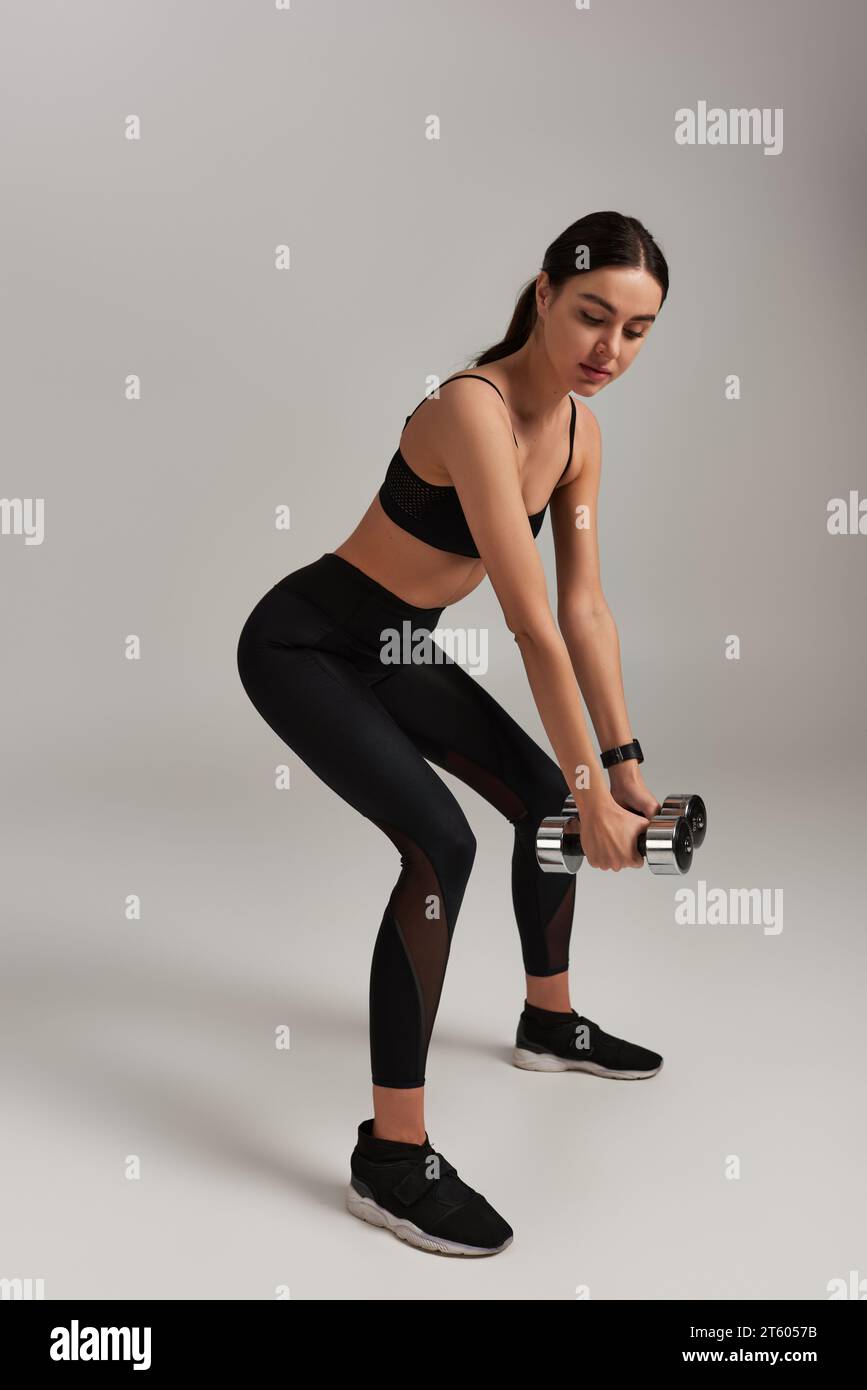 longitud completa de mujer poderosa en pesas de elevación de desgaste activo mientras hace ejercicio en el fondo gris Foto de stock