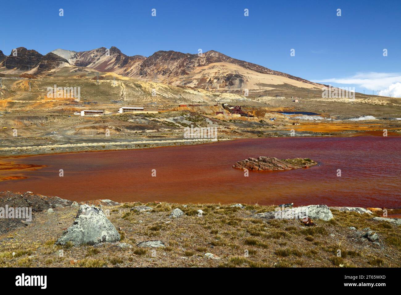 Vista a través del lago contaminado por el drenaje ácido de la mina y los residuos industriales de la mina de estaño cercana en Milluni (parte de la cual se puede ver en la orilla lejana y ladera de la montaña), cerca de La Paz, Bolivia. Las montañas son las estribaciones del macizo del monte Chacaltaya. Foto de stock
