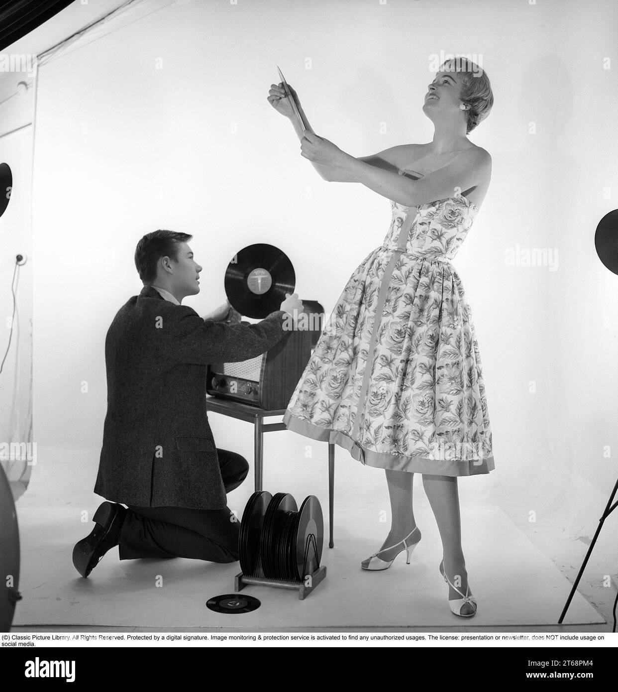 Gramófono en la década de 1950 Un hombre está arrodillado por un conjunto de recitación combinado y un gabinete de gramófono donde ha abierto la cubierta del gramófono y tiene un registro en su mano. Ella está vestida con un vestido típico de los años 1950 que se ve feliz. Suecia 1959. Kristoffersson ref CD65-2 Foto de stock
