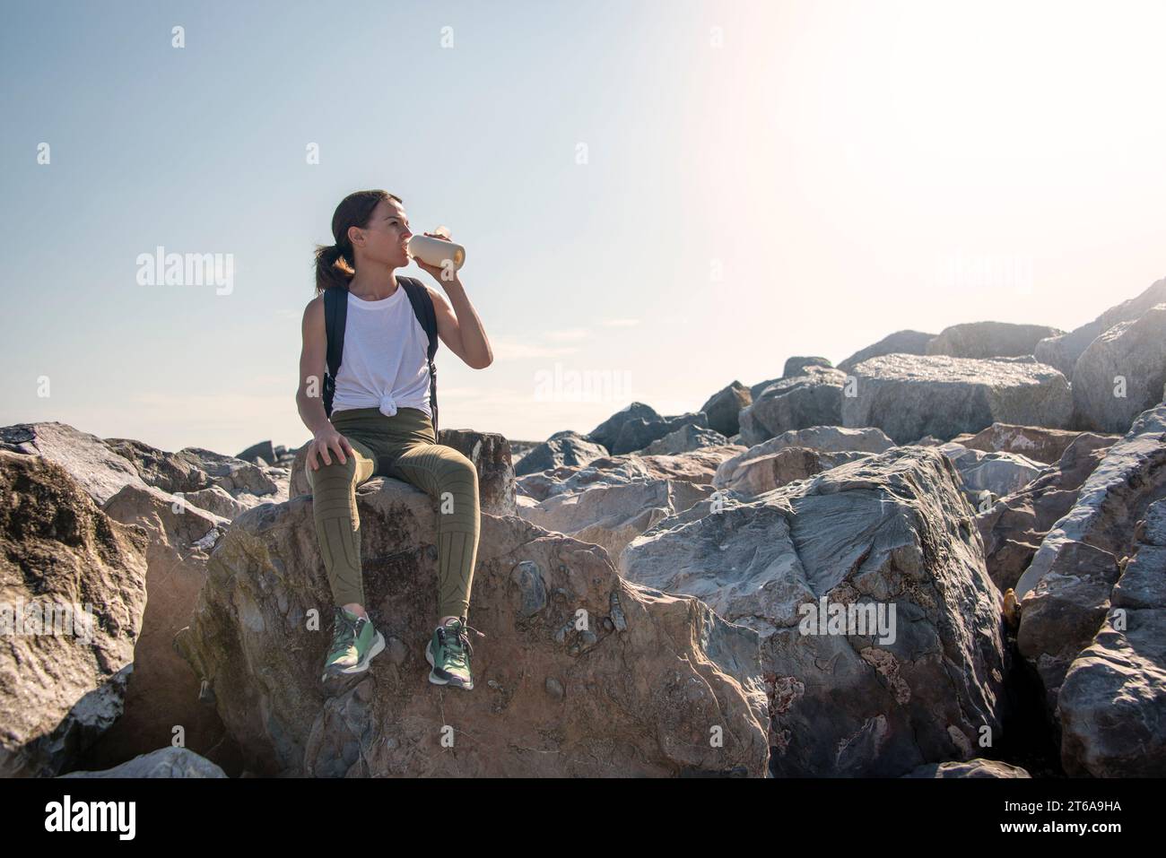 Una mujer excursionista se sienta en la roca descansando al sol, bebiendo agua de una botella Foto de stock