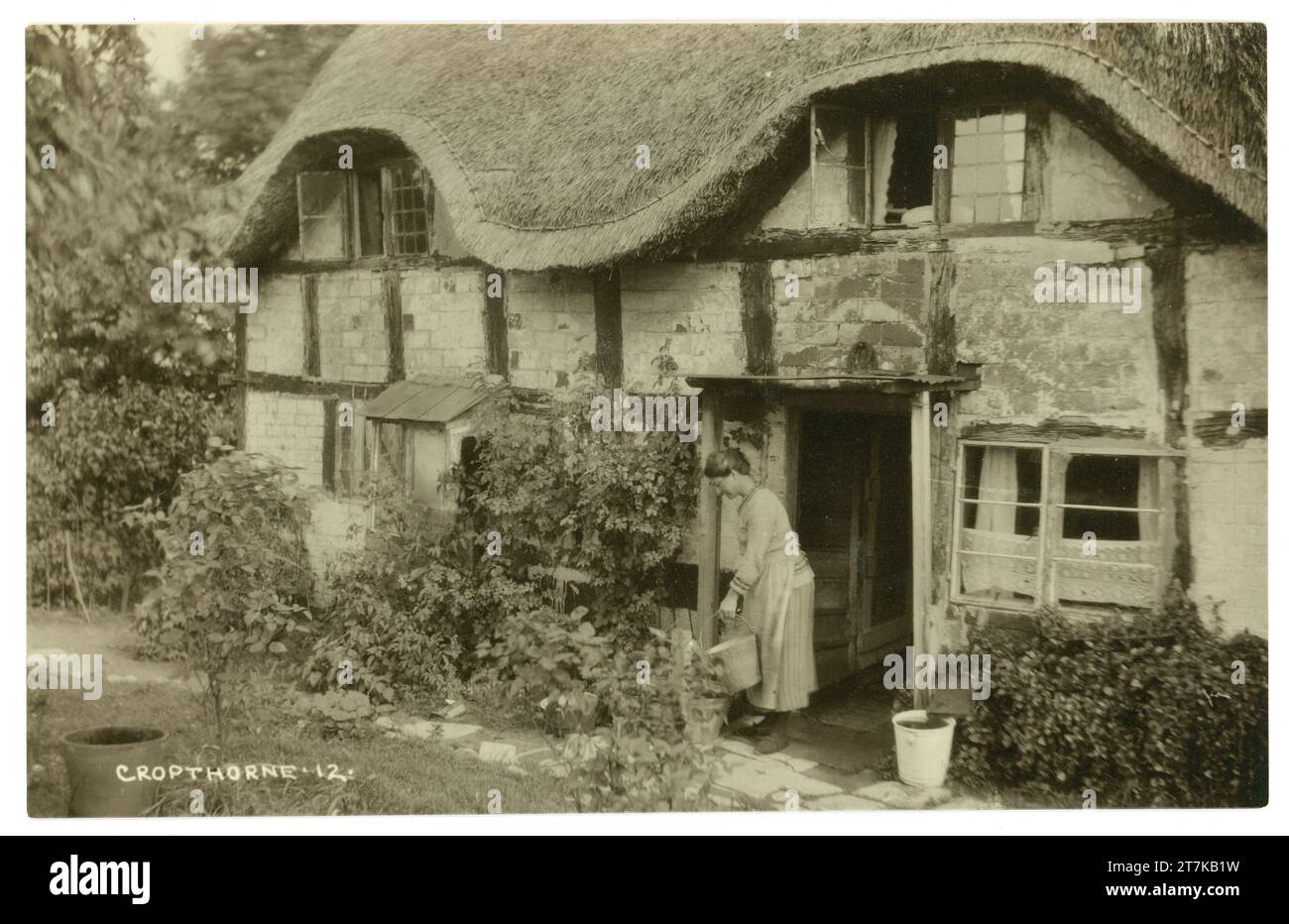 Postal original de la década de 1900 de una dama con balde / cubo fuera de su encantadora casa de campo de paja típica inglesa de entramado de madera, arquitectura vernácula, que data del período medieval, Cropthorne, Worcestershire. Inglaterra, Reino Unido Circa 1917 Foto de stock