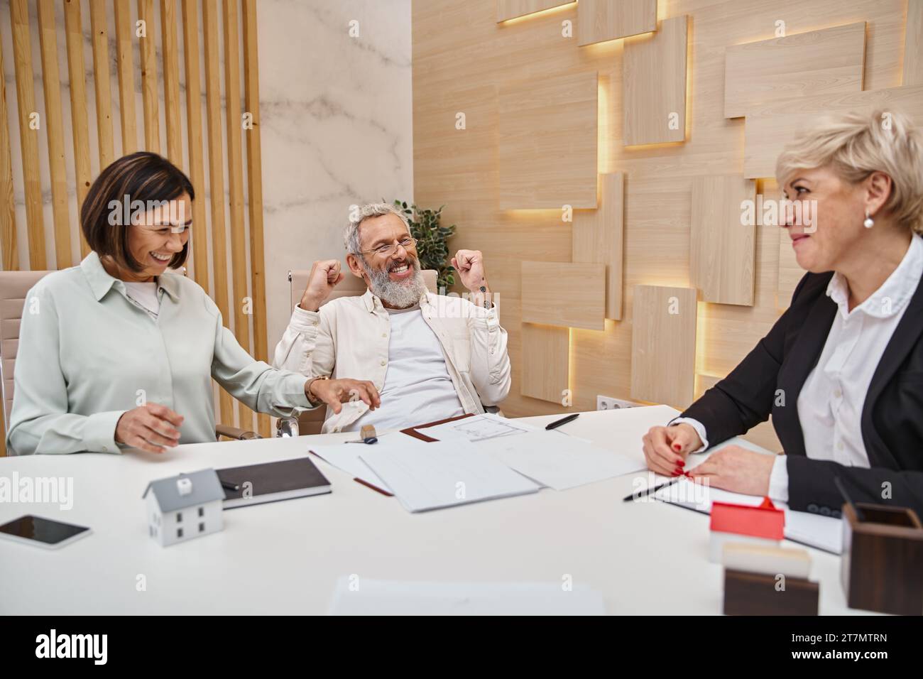 mujer feliz tomando la llave de la casa nueva cerca excitado marido de mediana edad y agente de bienes raíces en la oficina Foto de stock