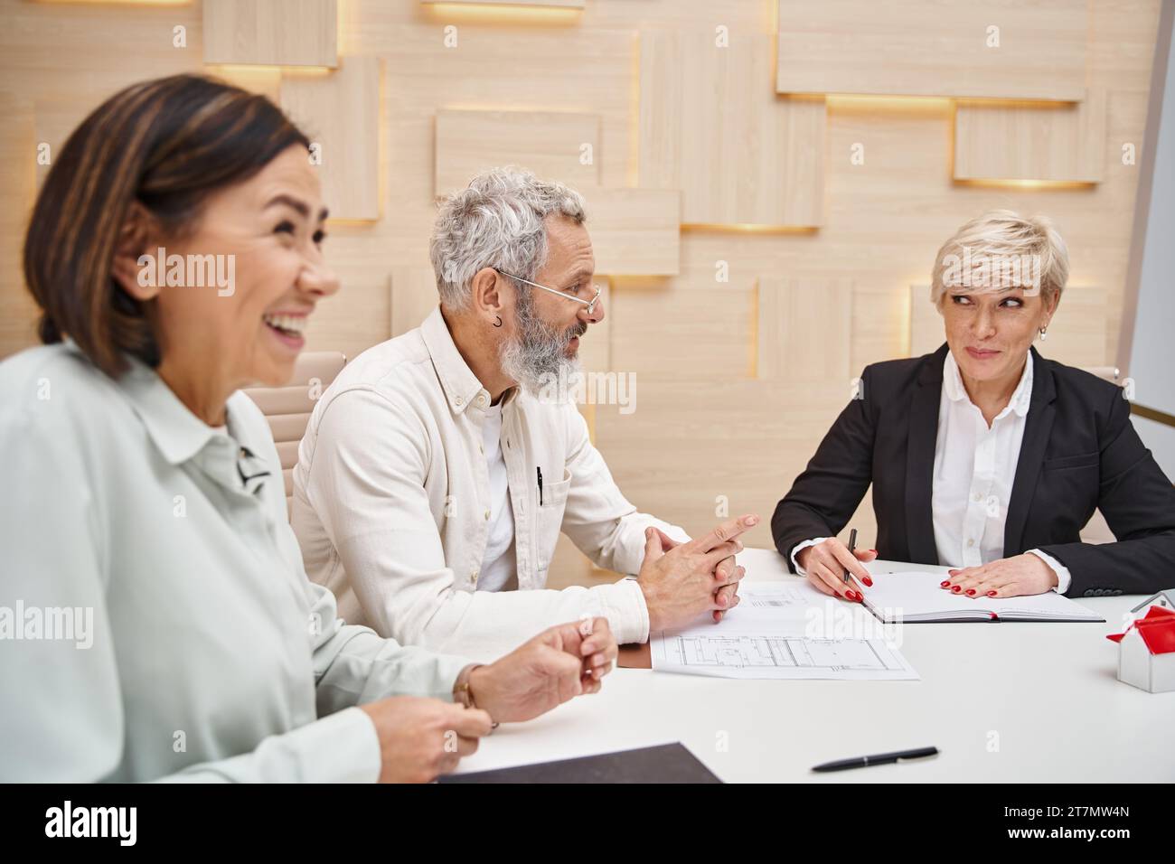 hombre de mediana edad feliz sonriendo cerca de la esposa y agente inmobiliario femenino mientras se sienta cerca del plano del nuevo hogar Foto de stock