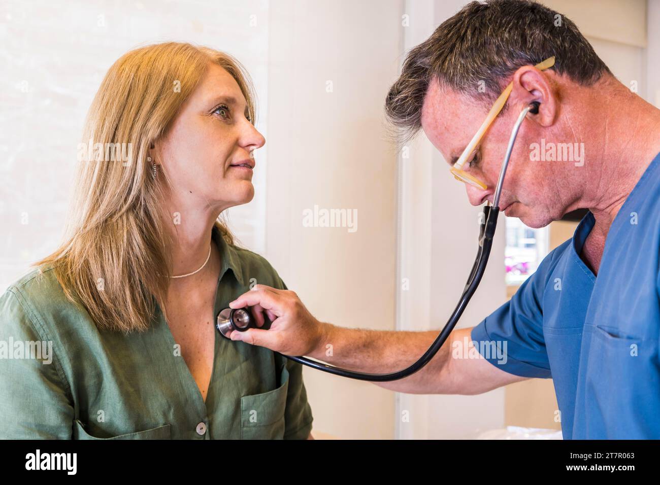 Un médico de mediana edad que llevaba ropa de exfoliación revisando el latido del corazón de su paciente con estetoscopio Foto de stock