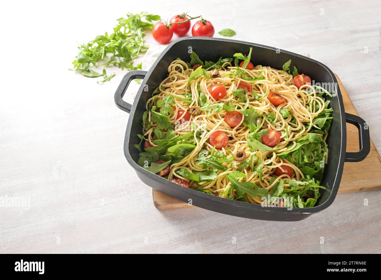 Espaguetis con tomates, rúcula y aceitunas en un gran tazón negro sobre una mesa pintada de blanco, comida vegetariana mediterránea casera, espacio de copia, seleccionar Foto de stock