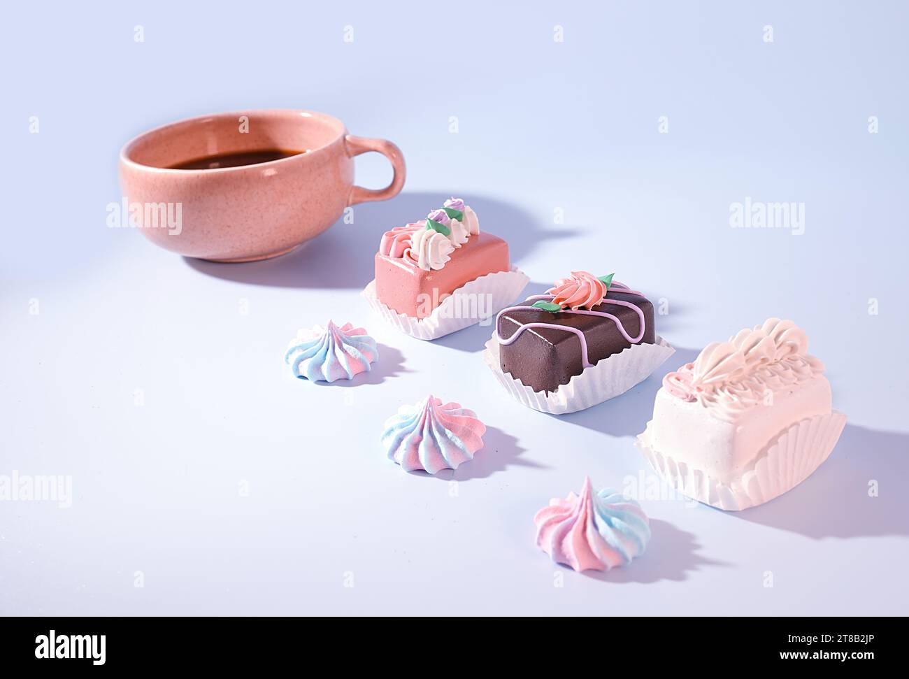Café y Confecciones: Una taza rosada de café con mini Petite cuatro pasteles decorados y merengue caramelo sobre fondo azul, golosinas de Pascua, panadería Concep Foto de stock
