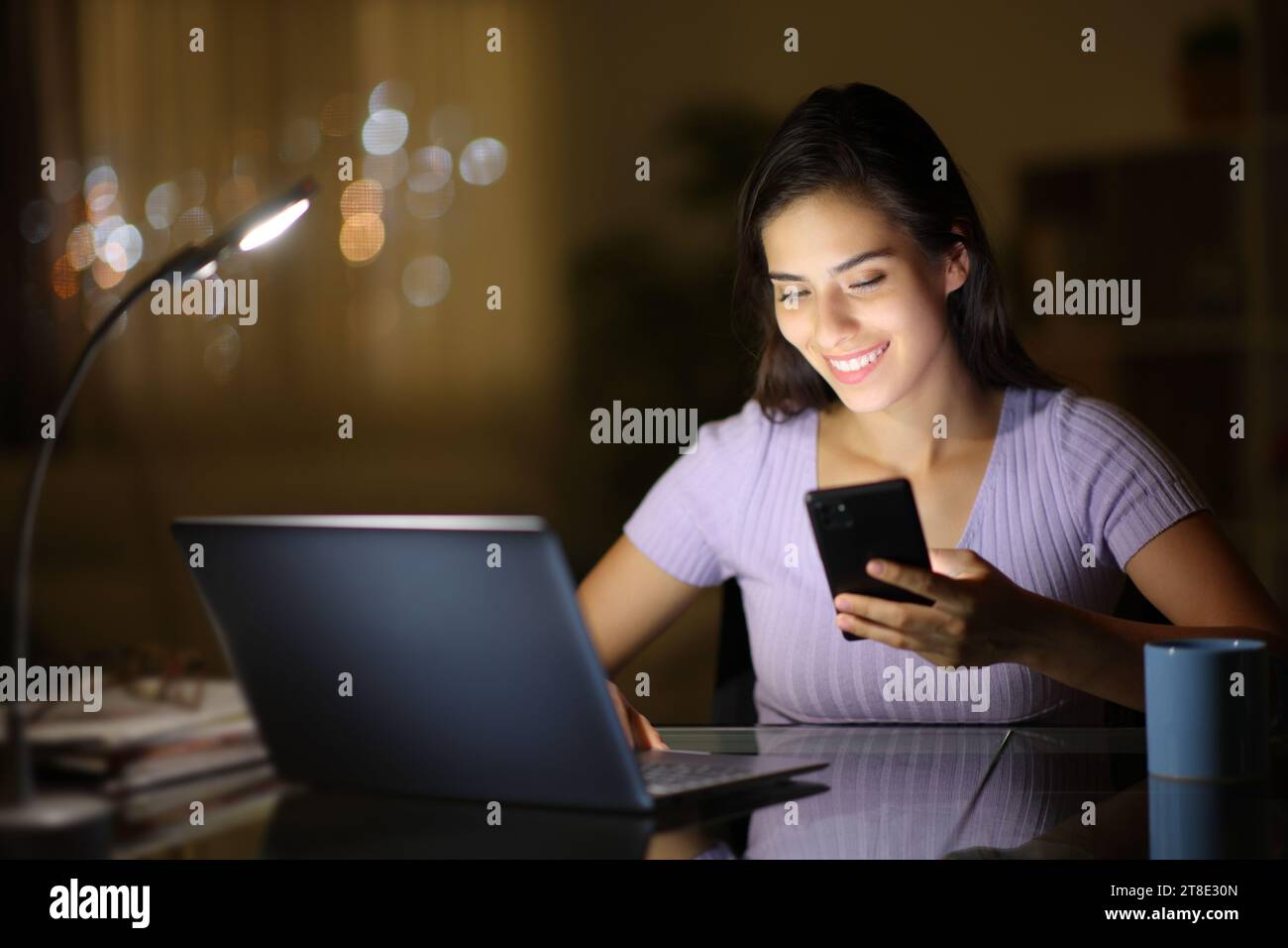 Mujer feliz que usa el ordenador portátil y el teléfono que comprueba en la noche en casa Foto de stock