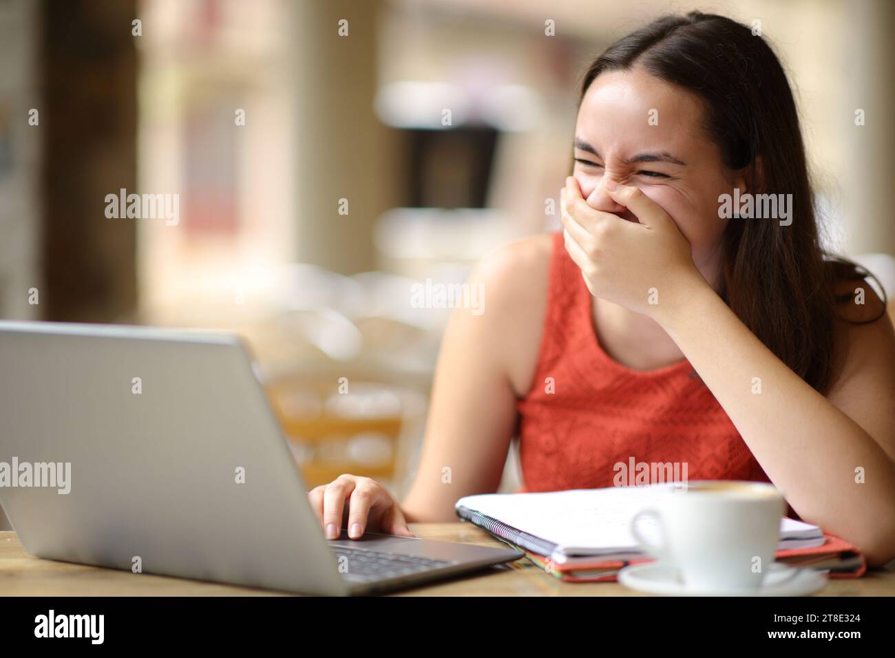 Estudiante riendo e-learning comprobando el ordenador portátil en una terraza de bar Foto de stock
