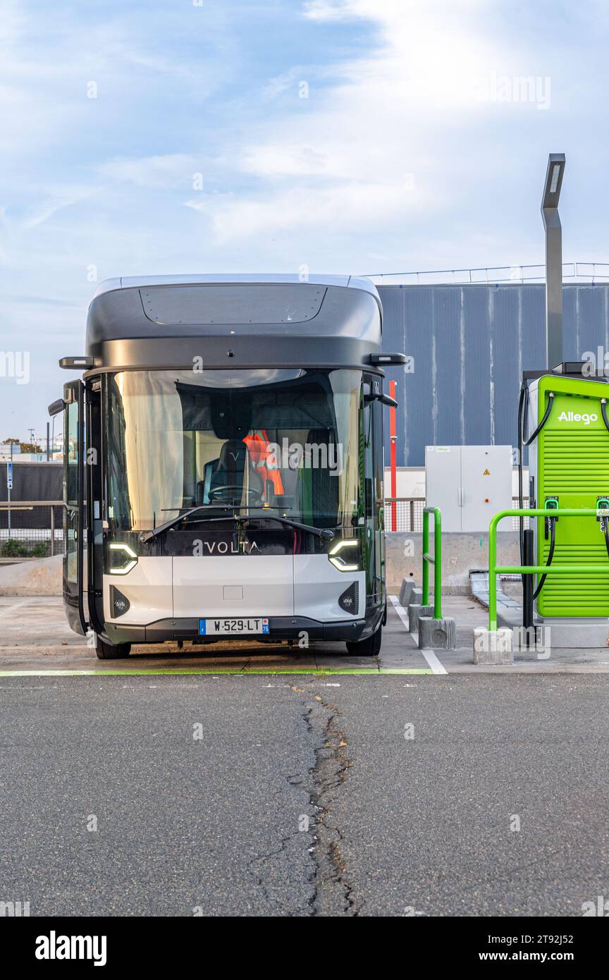 Camión eléctrico Volta totalmente eléctrico estacionado en una carretera, con un cielo azul vivo por encima. Junto a ella hay una estación de carga verde, que simboliza la sostenibilidad. Foto de stock