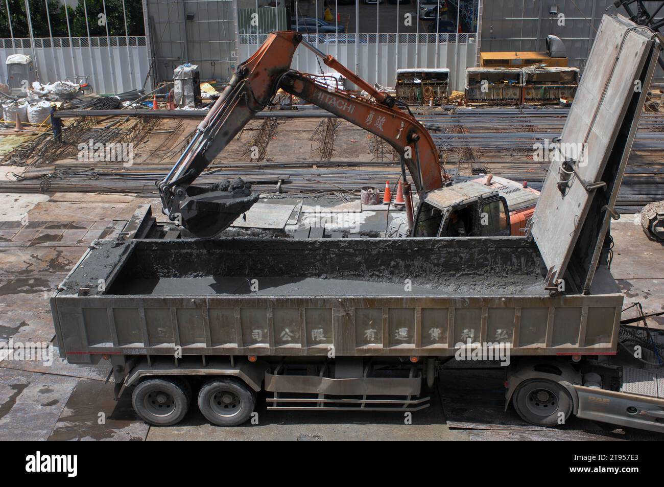 Excavadora que trabaja en una obra durante la construcción de un nuevo bloque de oficinas, cargando un camión con tierra fangosa en preparación para la colocación de cimientos, Foto de stock