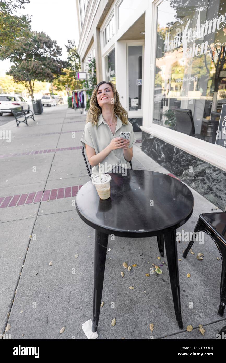 Hermosa mujer joven sentada con un café helado en una acera Café Downtown Foto de stock