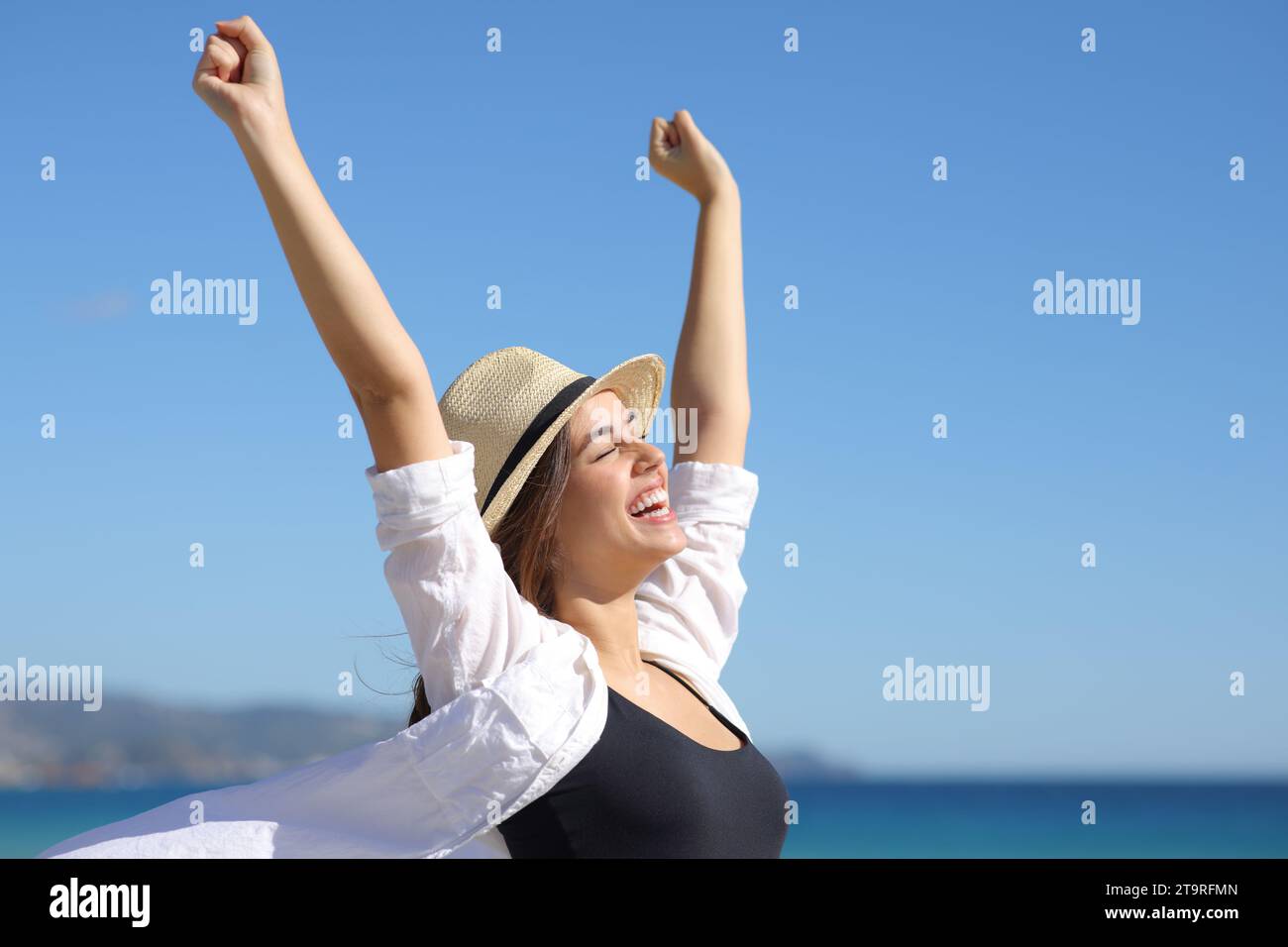 Mujer emocionada con sombrero celebrando un buen día levantando los brazos en la playa un día soleado Foto de stock