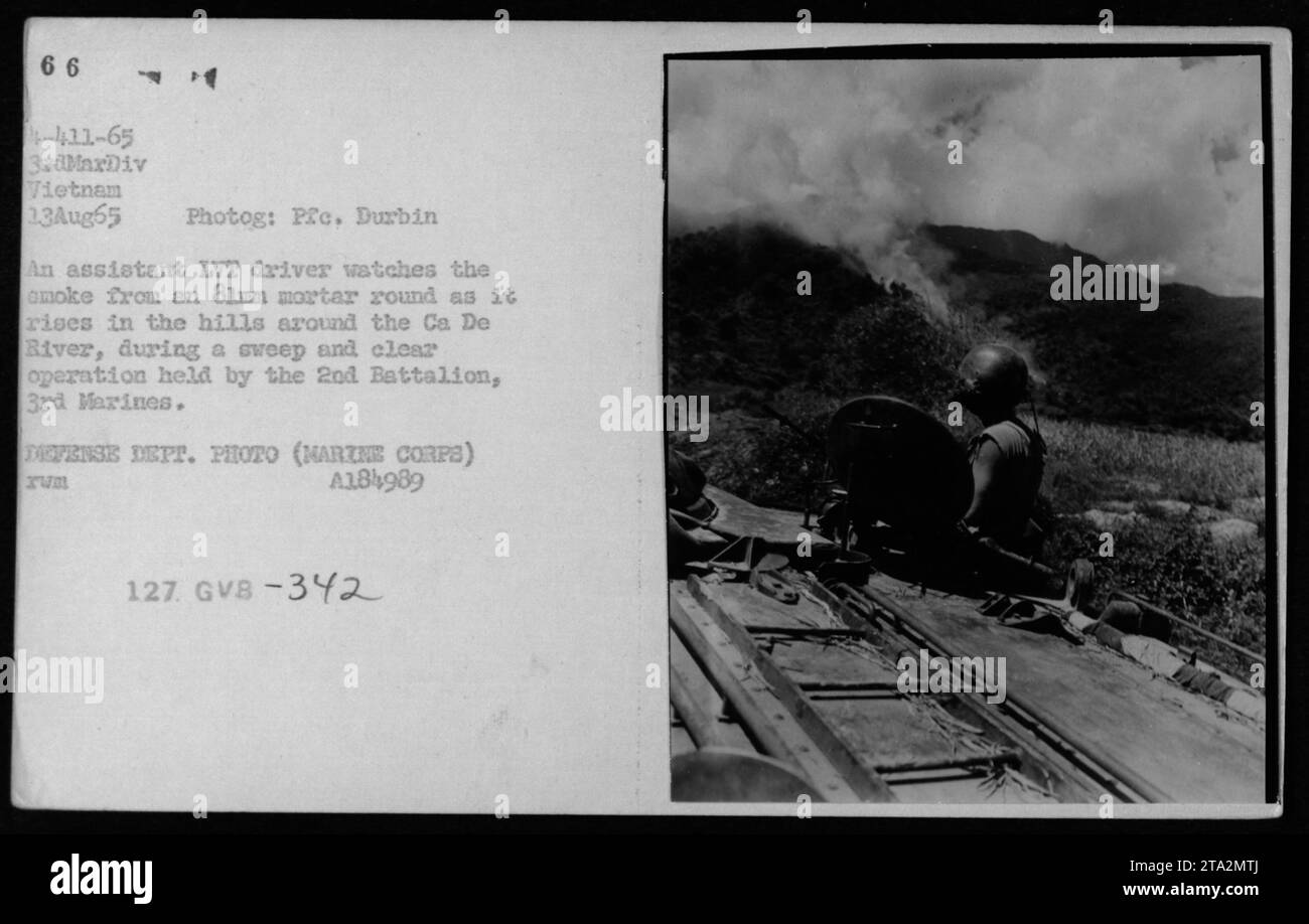 Durante una operación de barrido y despejado en Vietnam, un conductor asistente de vehículo de aterrizaje rastreado (LVT) mira como el humo de una ronda de mortero de 81 mm se eleva en las colinas que rodean el río Ca De. La fotografía fue tomada el 13 de agosto de 1965 por el PFC Durbin de la 3ra División de Marines. Foto de stock