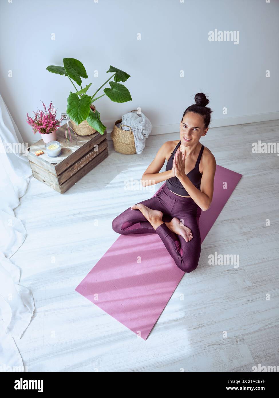 Alto ángulo de mujer sonriente en ropa deportiva sentado en esterilla de yoga púrpura en pose de loto con gesto de namaste y mirando a la cámara en casa Foto de stock