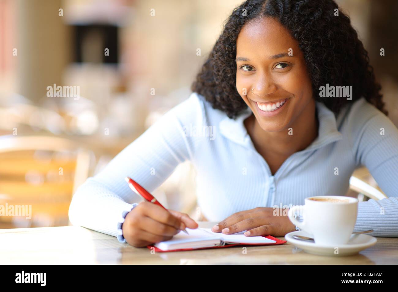 Mujer negra feliz te mira escribiendo en agenda de papel en una terraza de restaurante Foto de stock