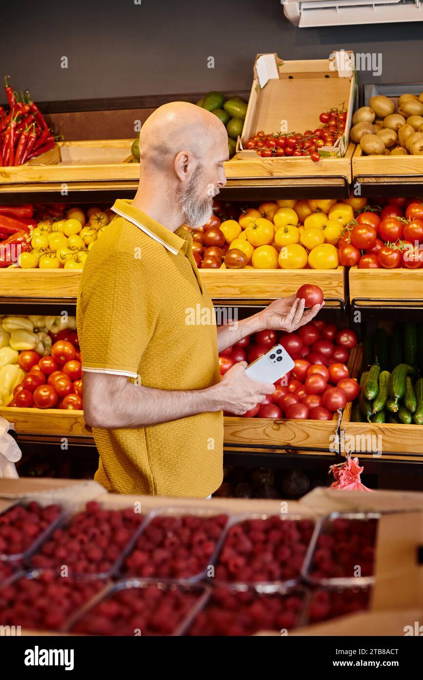 cliente maduro barbudo gris que sostiene el teléfono móvil y mira el tomate jugoso fresco en la tienda de comestibles Foto de stock