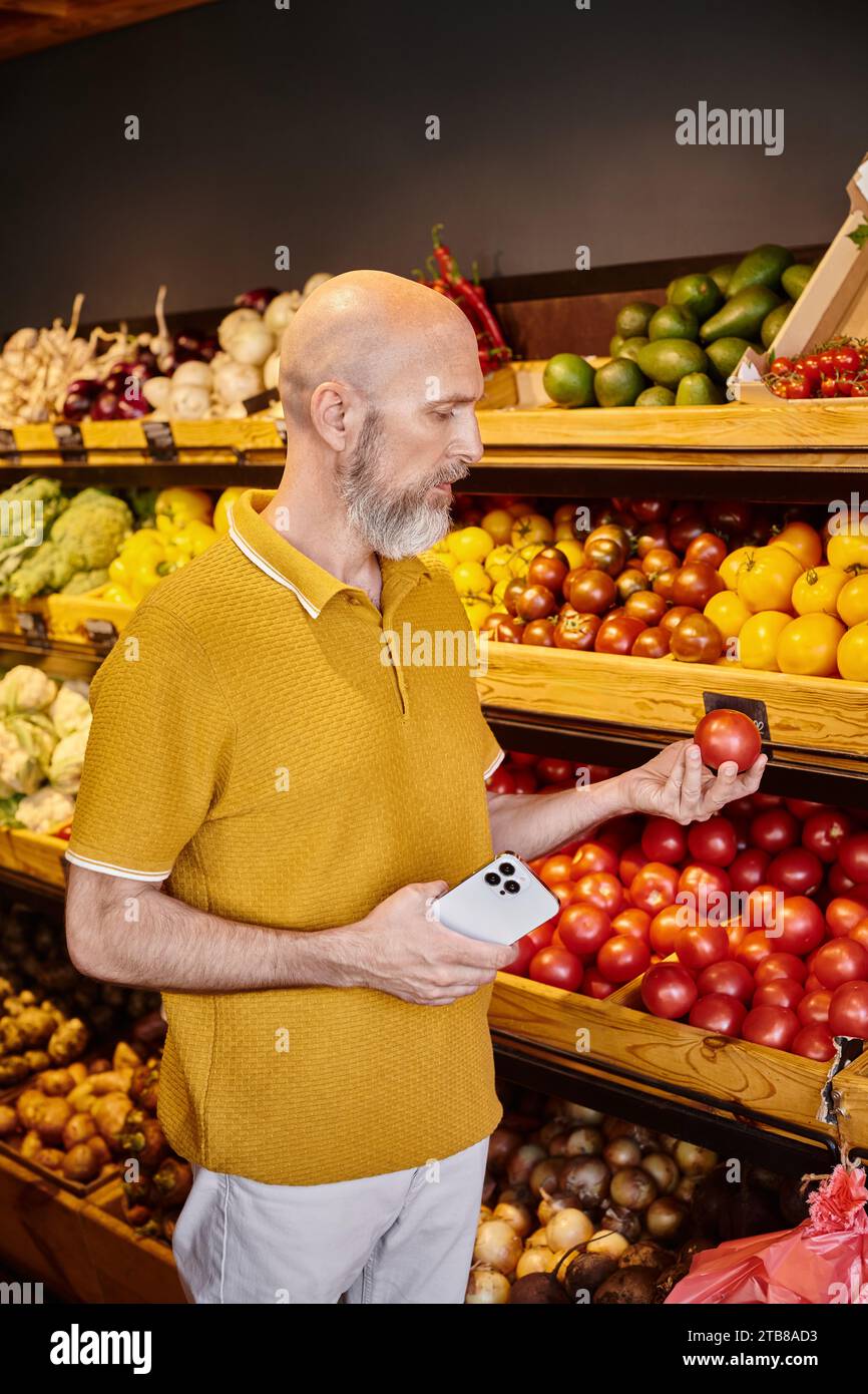 hombre maduro de buen aspecto con barba sosteniendo el teléfono móvil y mirando el tomate fresco en la tienda de comestibles Foto de stock