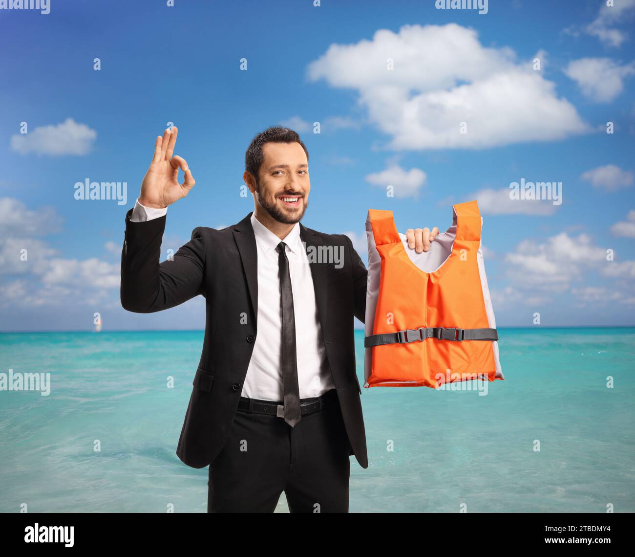 Hombre en un traje y corbata sosteniendo un chaleco salvavidas junto al mar y gestando una señal ok Foto de stock