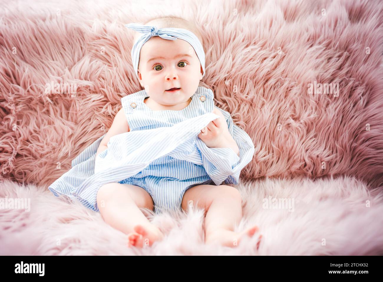 Lindo niño sonriente en un sofá de color rosa. Los primeros dientes. Salud 6 meses bebé chica sentada. Vestido azul Foto de stock