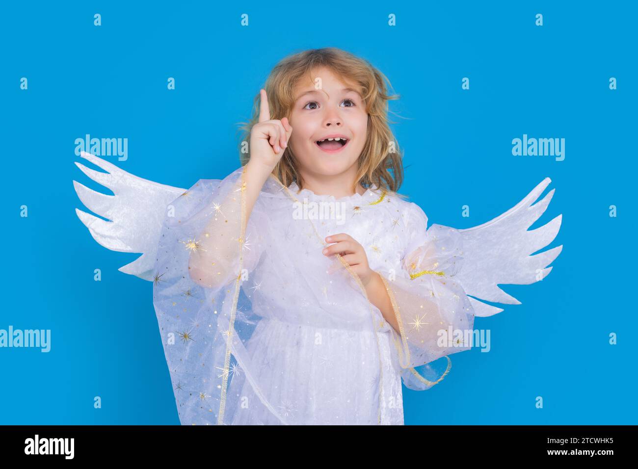 Niño ángel apuntando hacia arriba, gesto de punto. Día de San Valentín. Niño lindo rubio con alas de ángel sobre un fondo de estudio azul. Niño ángel feliz. Foto de stock