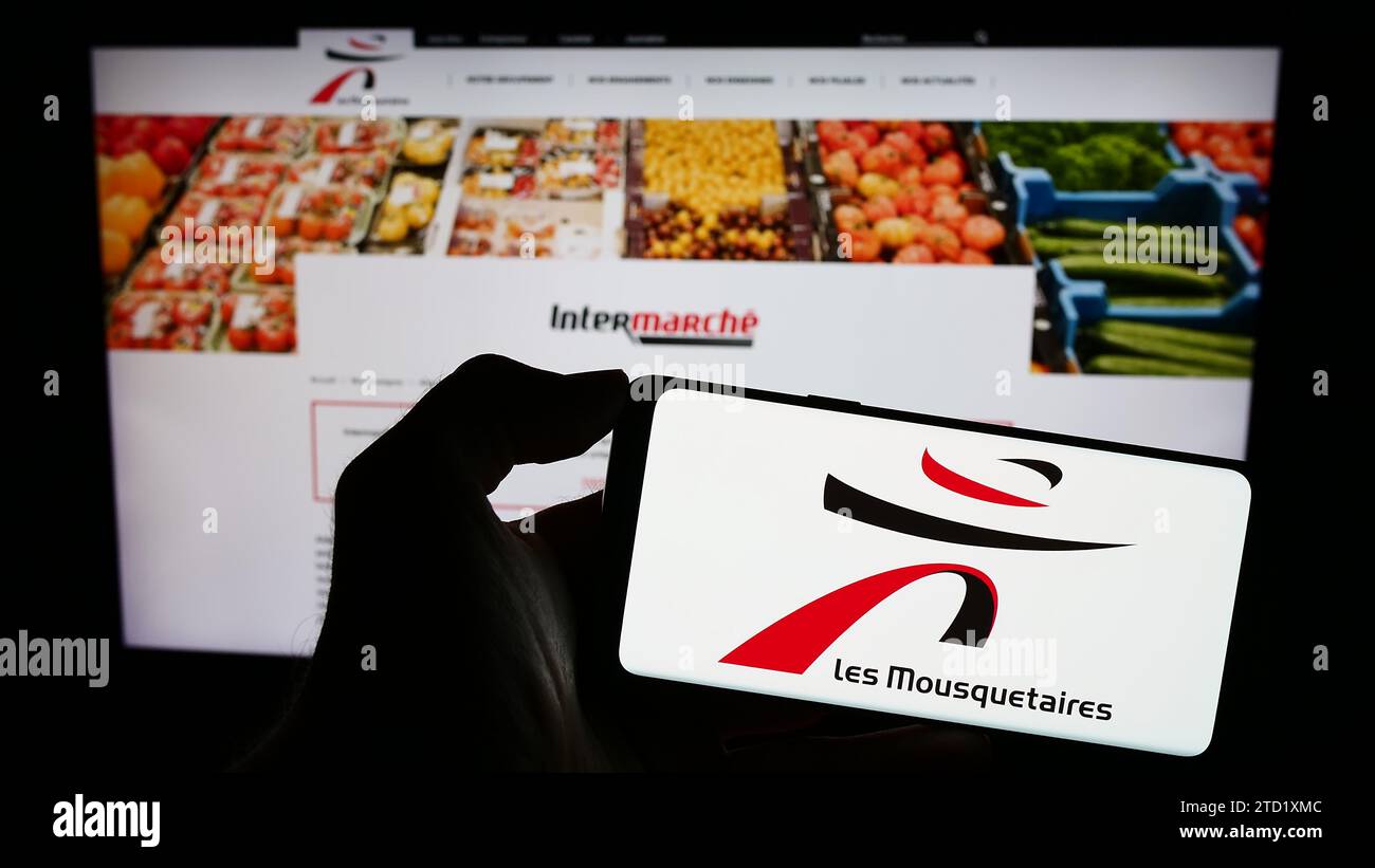 Persona que sostiene el teléfono móvil con el logotipo de la empresa francesa ITM Entreprises SA (Les Mousquetaires) delante de la página web. Enfoque en la pantalla del teléfono. Foto de stock