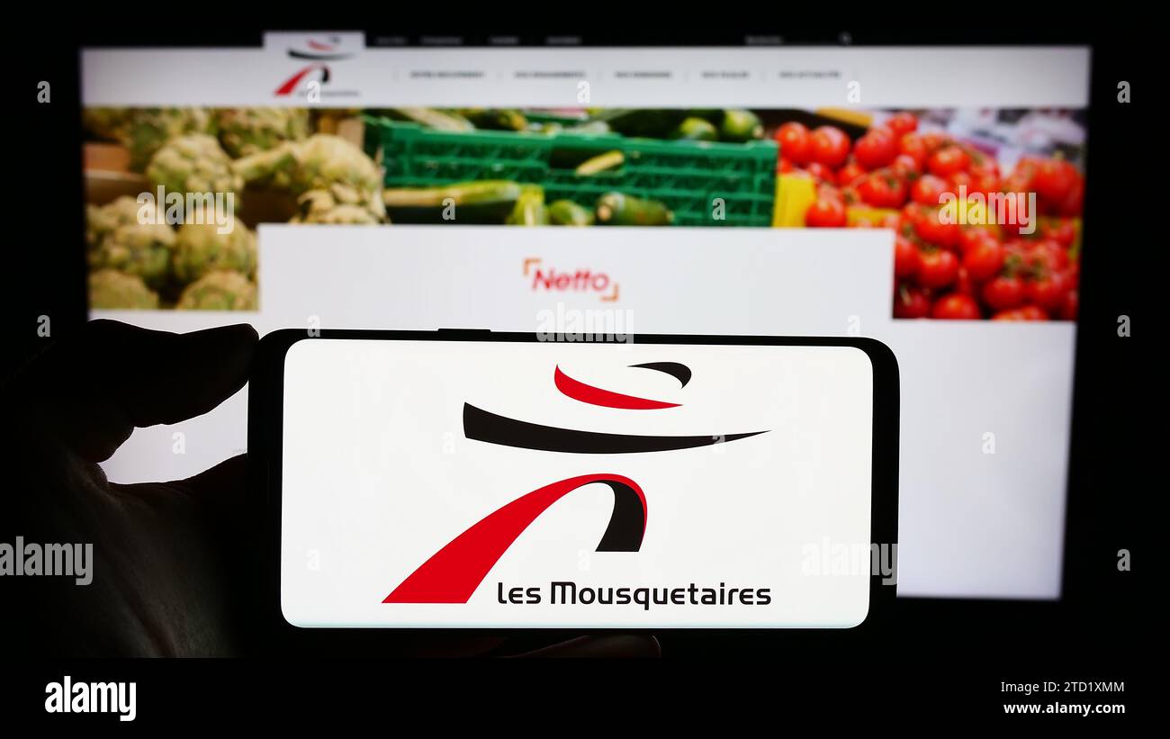 Persona titular del teléfono móvil con el logotipo de la empresa francesa ITM Entreprises S.A. (Les Mousquetaires) delante de la página web. Enfoque en la pantalla del teléfono. Foto de stock