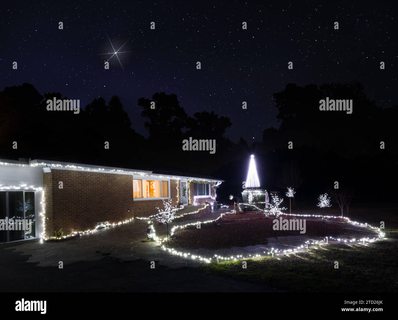 Pequeña casa de granja con todas las luces blancas que decoran el patio durante las vacaciones con una estrella brillante que se eleva en el cielo detrás en la víspera de Navidad con la t Foto de stock