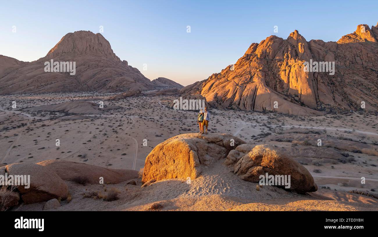 Pareja en una montaña al atardecer, cumbre del Gran Spitzkoppe y Pontoks en el fondo, rocas erosionadas, Parque Nacional Spitzkoppe, Erongo, Namibia Foto de stock