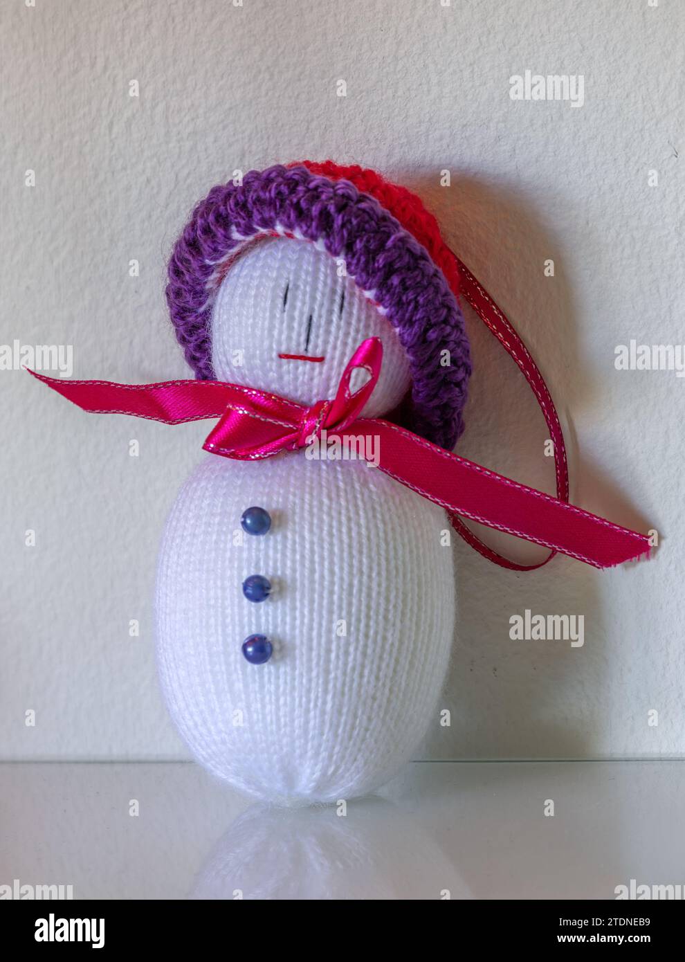 Primer plano de una muñeca de Navidad, hecha a mano en crochet, con botones, cinta y sombrero, de pie sobre una superficie de vidrio contra una pared blanca. Foto de stock