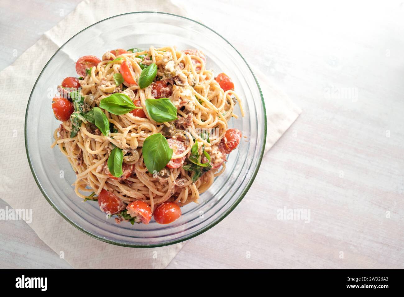 Ensalada fría de espaguetis con tomates, rúcula, mozzarella, aceitunas y un aderezo cremoso con adorno de albahaca en un tazón de vidrio sobre una mesa pintada de blanco, encima Foto de stock