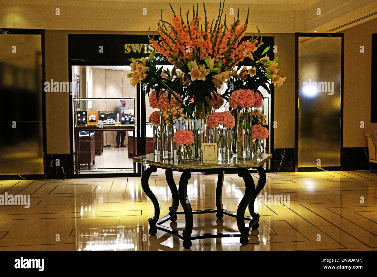Un elegante vestíbulo del hotel con una suntuosa exhibición floral que incluye hortensias, lilos y gladiolos en varios tonos de rosa con una iluminación ambiental suave Foto de stock