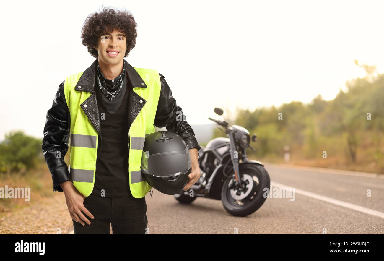 Hombre joven con una moto sosteniendo un casco y usando un chaleco de seguridad de tráfico en una carretera abierta Foto de stock