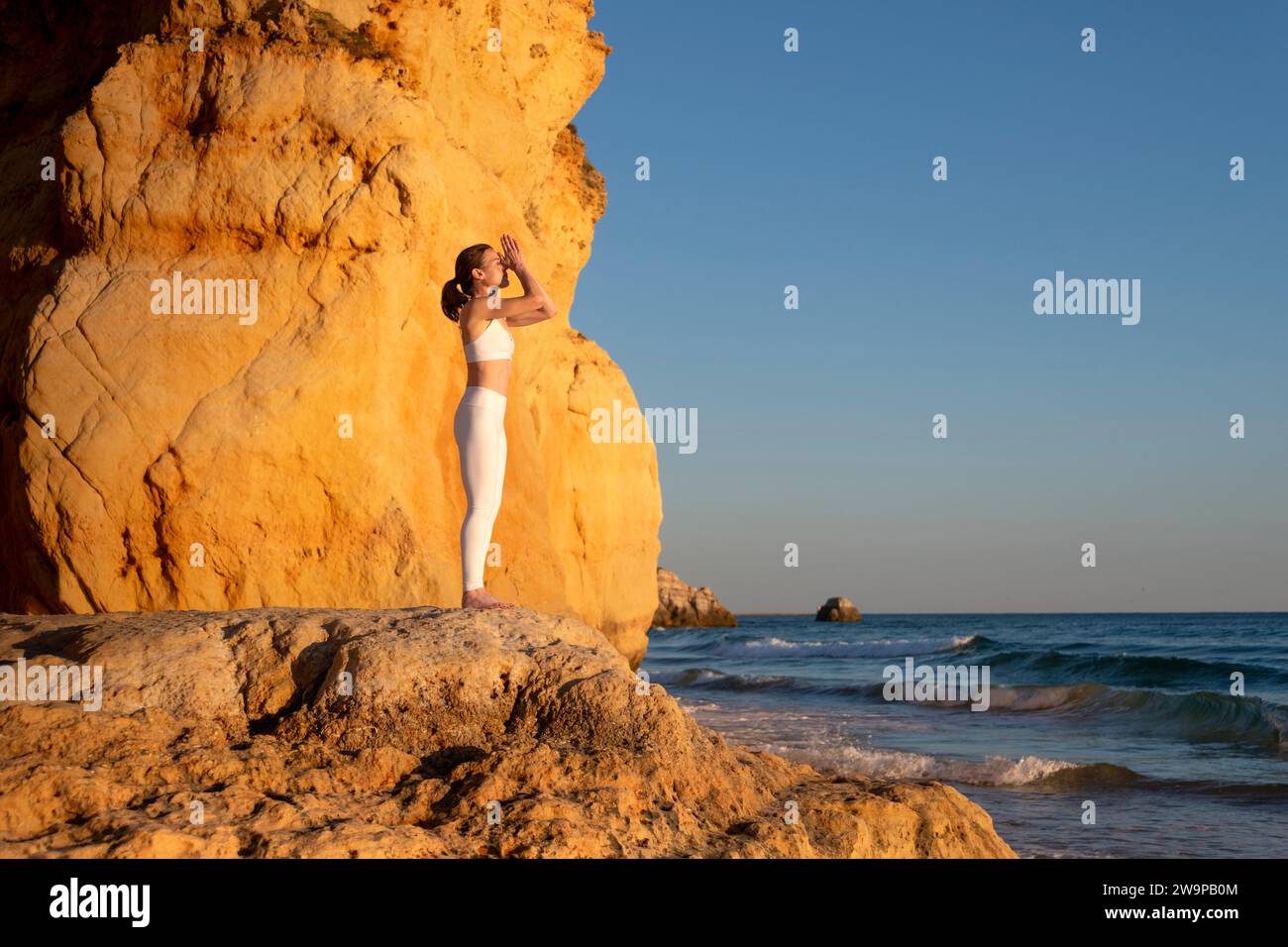 Mujer meditando y practicando yoga de pie en las rocas junto al mar en el sol de la tarde. Foto de stock