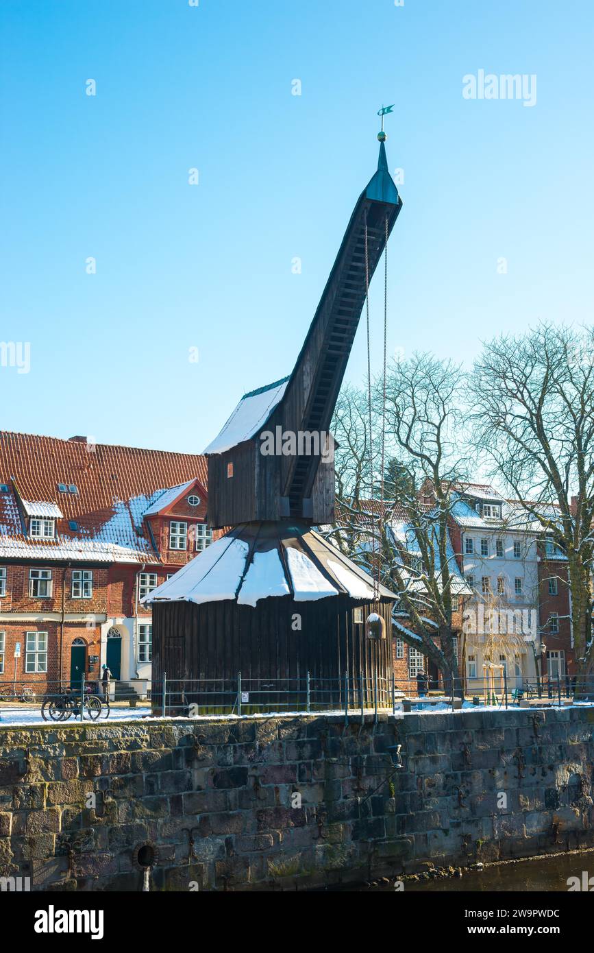 Antigua grúa en la ribera nevada del río Ilmenau frente a edificios históricos bajo un cielo azul claro, día soleado de invierno, árboles viejos, casco antiguo histórico Foto de stock