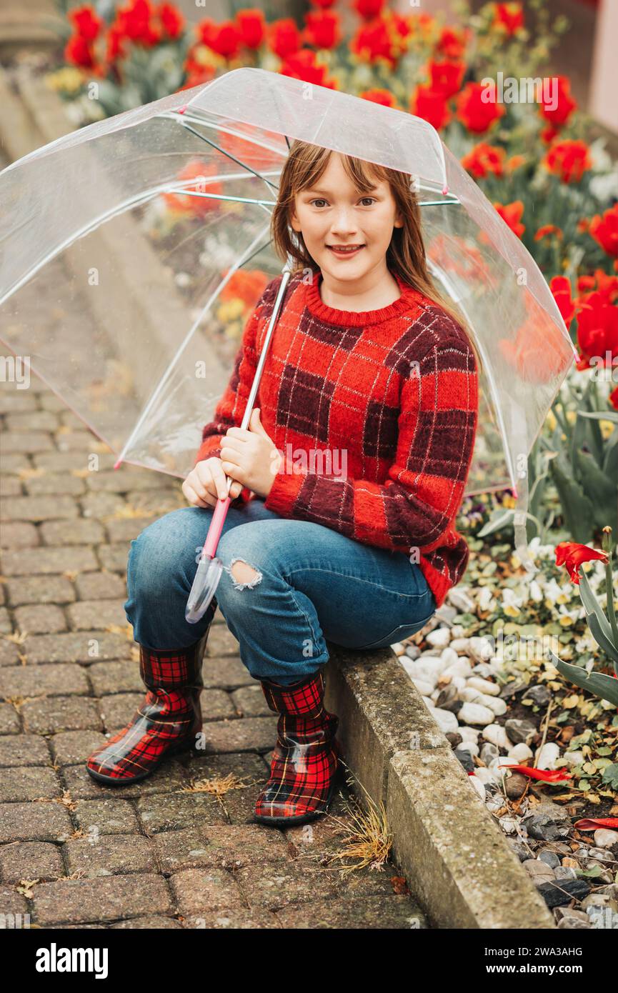 Retrato al aire libre de la primavera de la muchacha adorable del niño que sostiene el paraguas transparente, usando el jersey rojo de cuadros y las botas de la lluvia, niño que juega afuera en un lluvioso Foto de stock