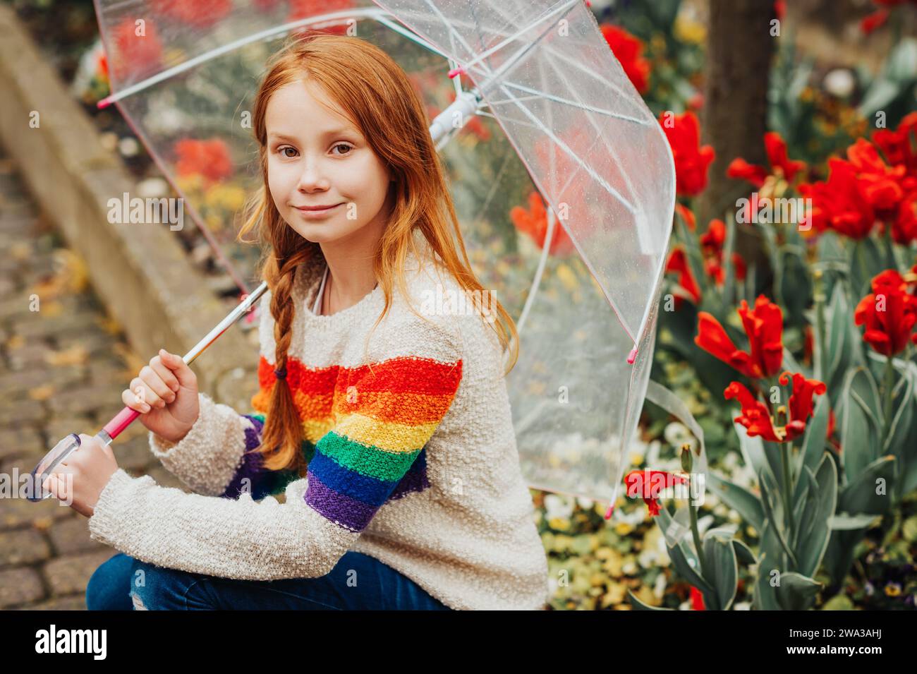 Retrato al aire libre de la primavera de la muchacha adorable pelirroja que sostiene el paraguas transparente, usando el jersey del arco iris, niño que juega afuera en un día lluvioso, f Foto de stock