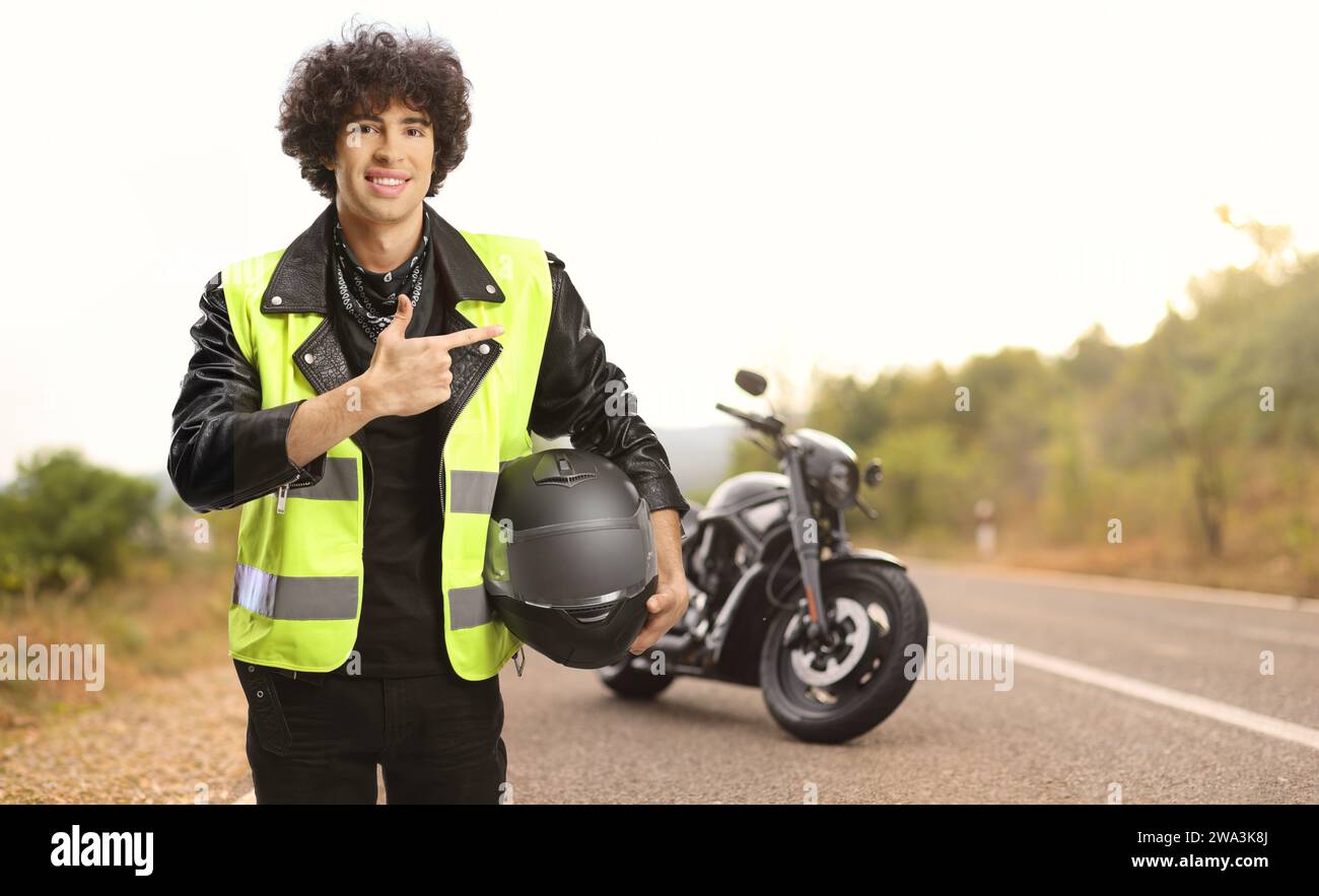 Joven apuntando a una moto, sosteniendo un casco y usando un chaleco de seguridad de tráfico en una carretera abierta Foto de stock
