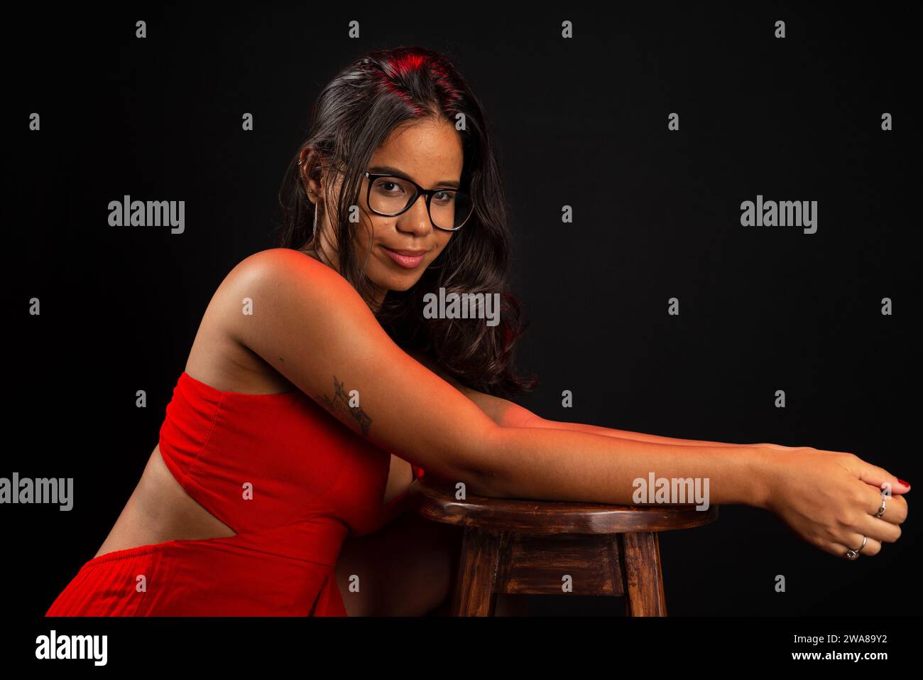 Retrato de la mujer joven hermosa que lleva gafas y en vestido rojo que se sienta posando para la foto. Aislado sobre fondo negro. Foto de stock