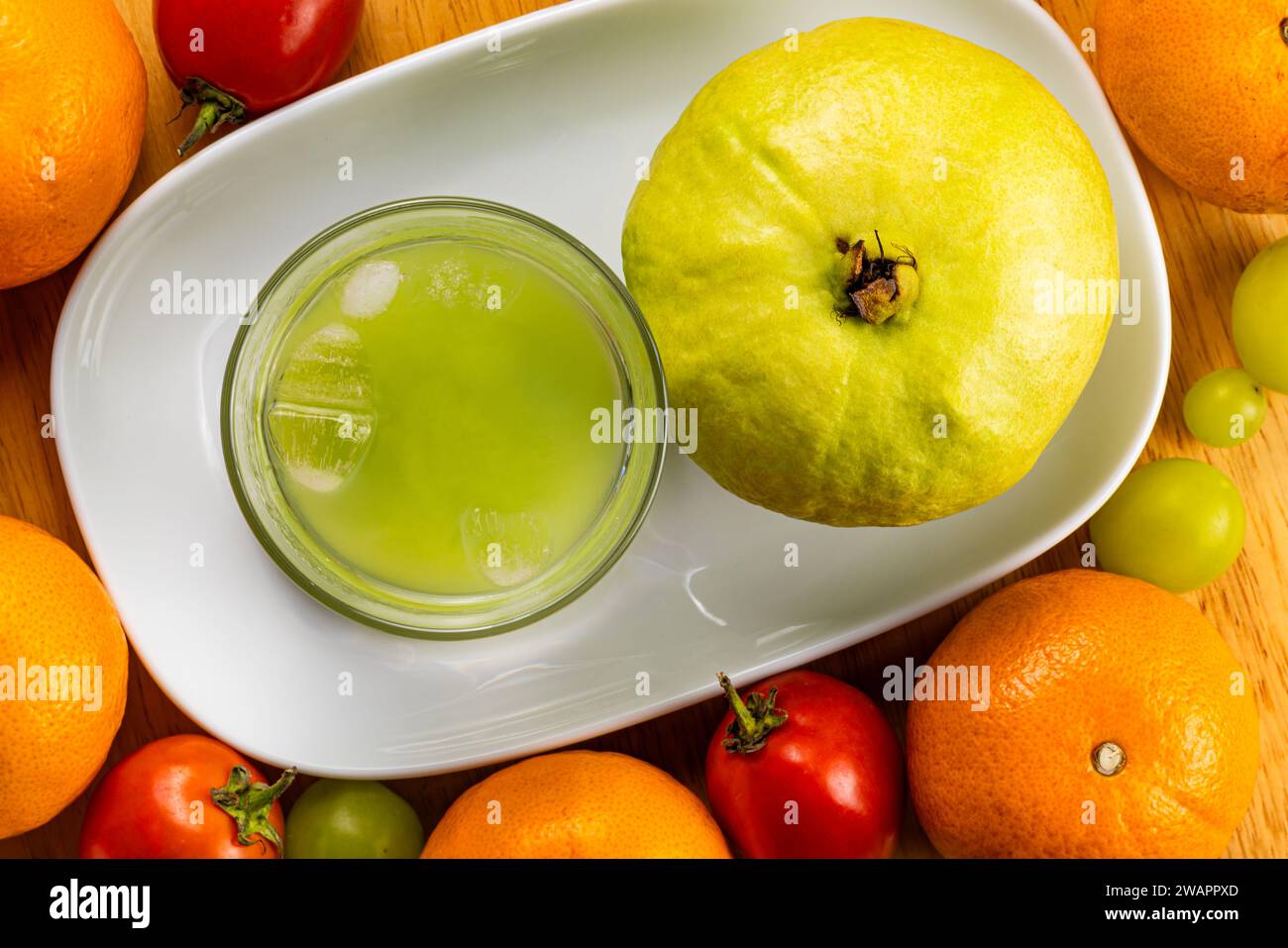 Vista superior de jugo de guayaba dulce fresco y frío en vidrio transparente y fruta de guayaba fresca madura en bandeja de cerámica blanca con tomate rojo, naranjas, uvas en woo Foto de stock